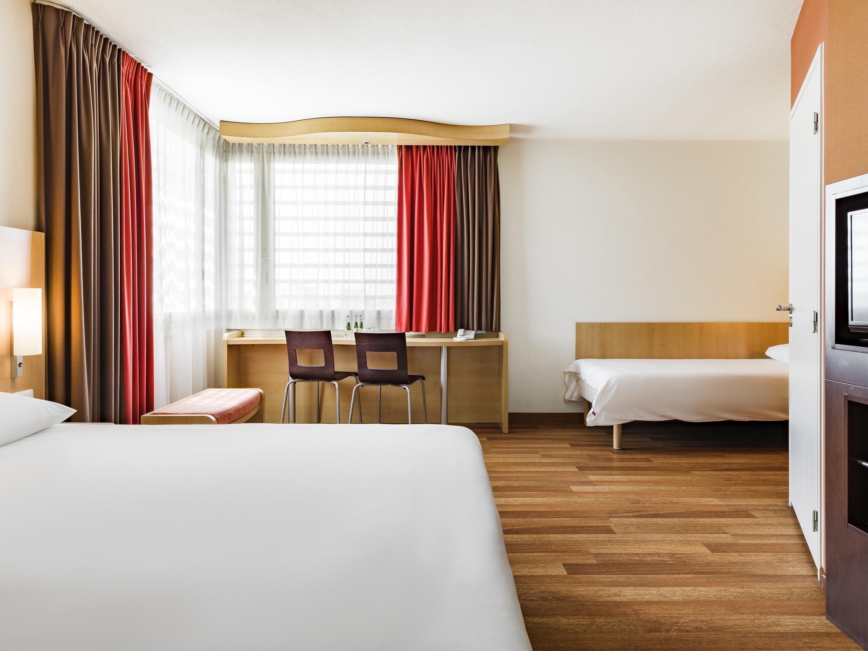 A hotel room with a bed, a desk with two chairs, a TV, and a window with curtains.