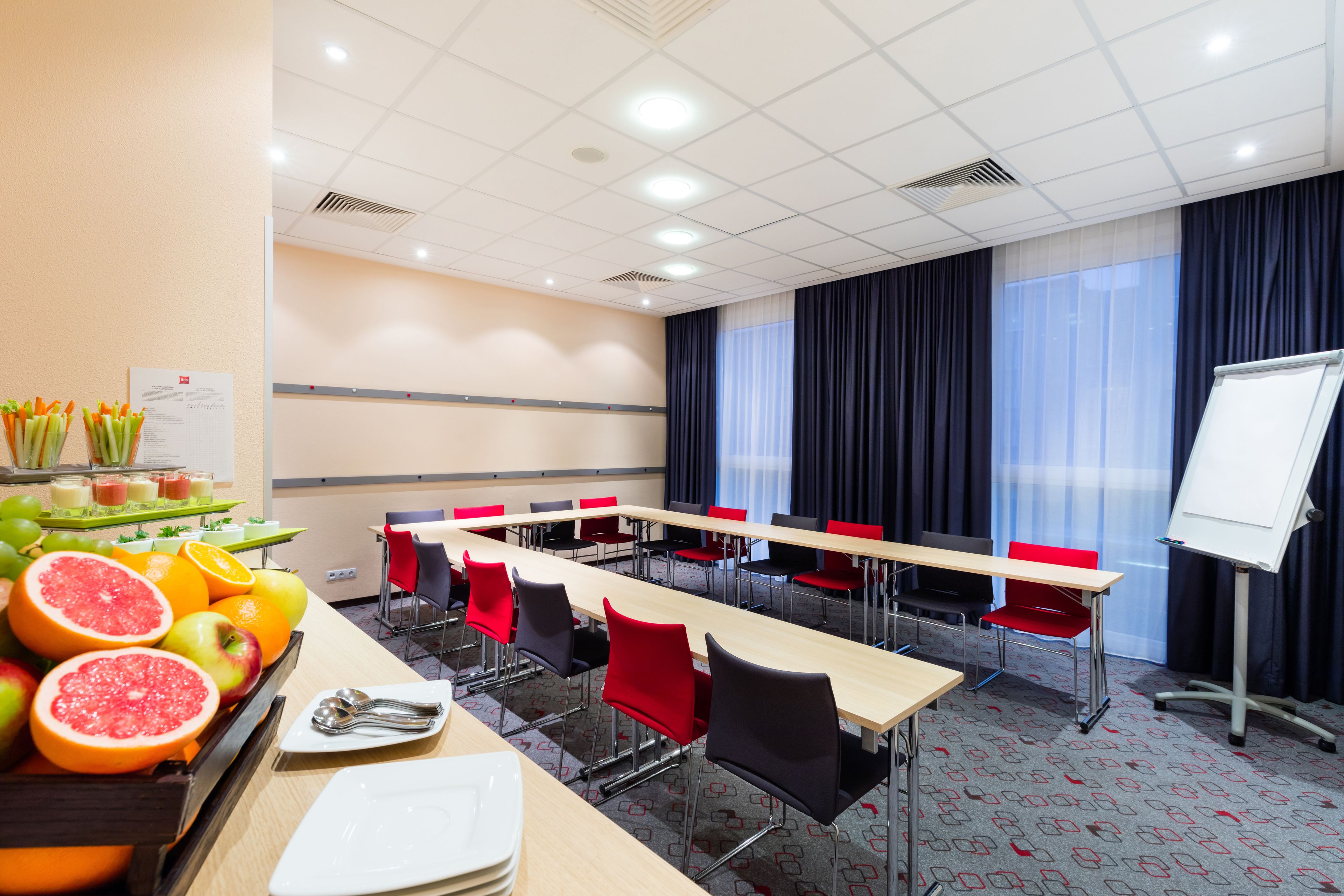 Ein Konferenzraum mit langen Tischen, roten Stühlen, einer Whiteboard und einer Obstauslage.