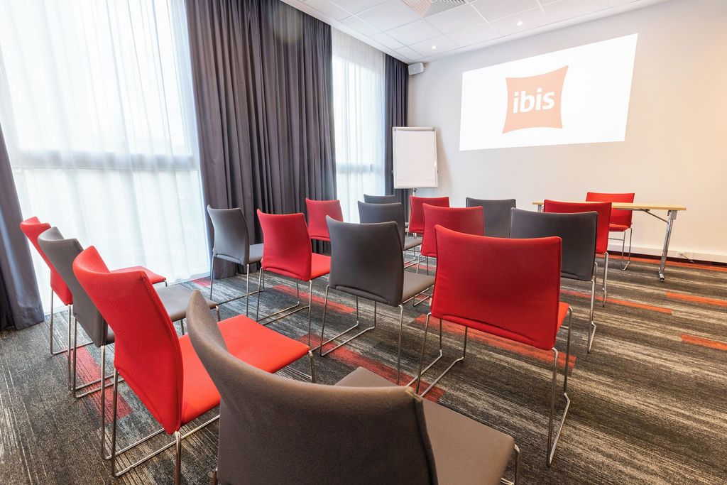Un salón de conferencias con una pantalla de proyector mostrando el logo de 'ibis', disposición de asientos y piso alfombrado.
