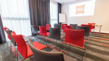 Un salón de conferencias con una pantalla de proyector mostrando el logo de 'ibis', disposición de asientos y piso alfombrado.