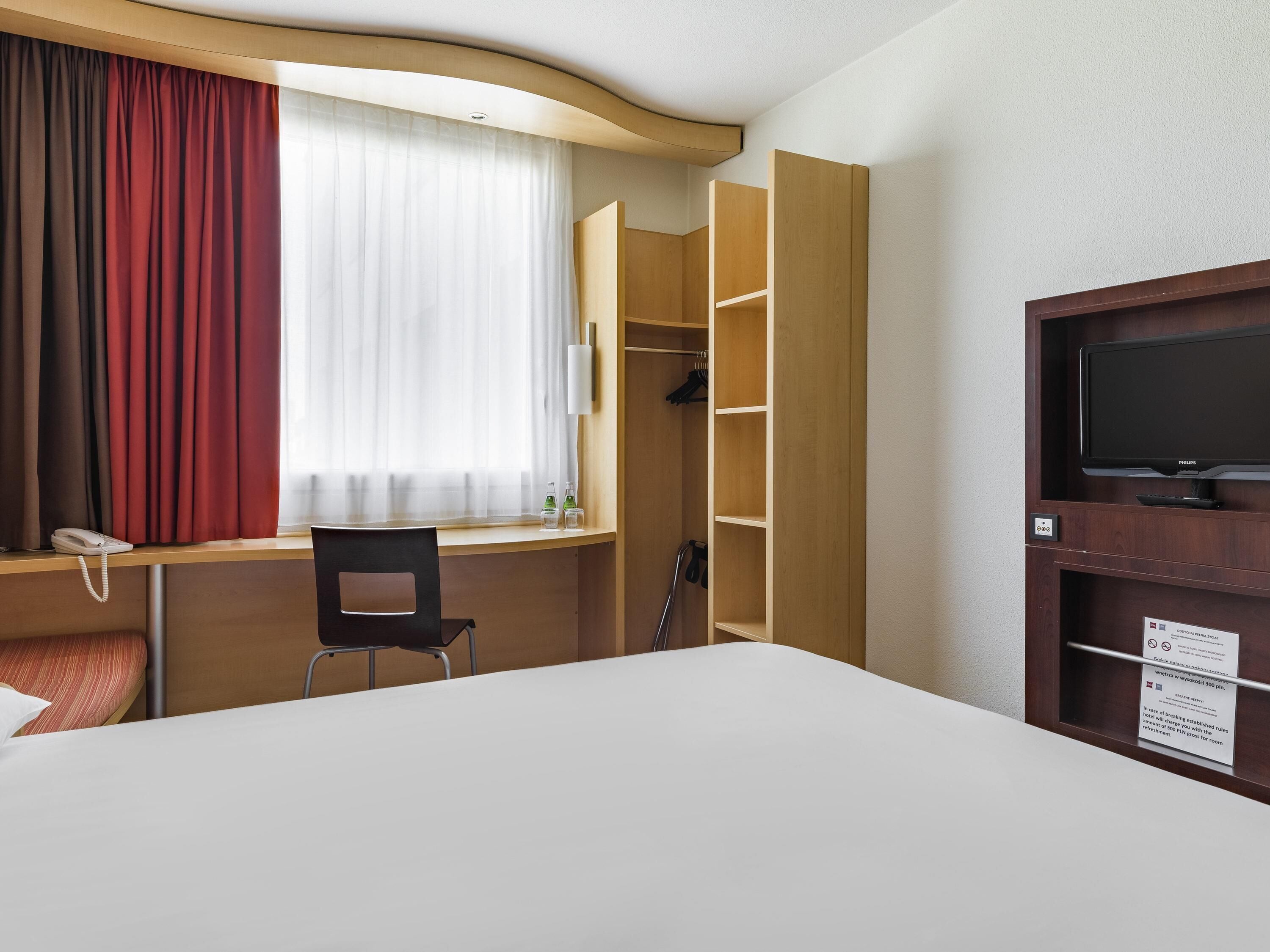 A hotel room with a bed, a desk with a chair, a television, a wardrobe, and a window with curtains.