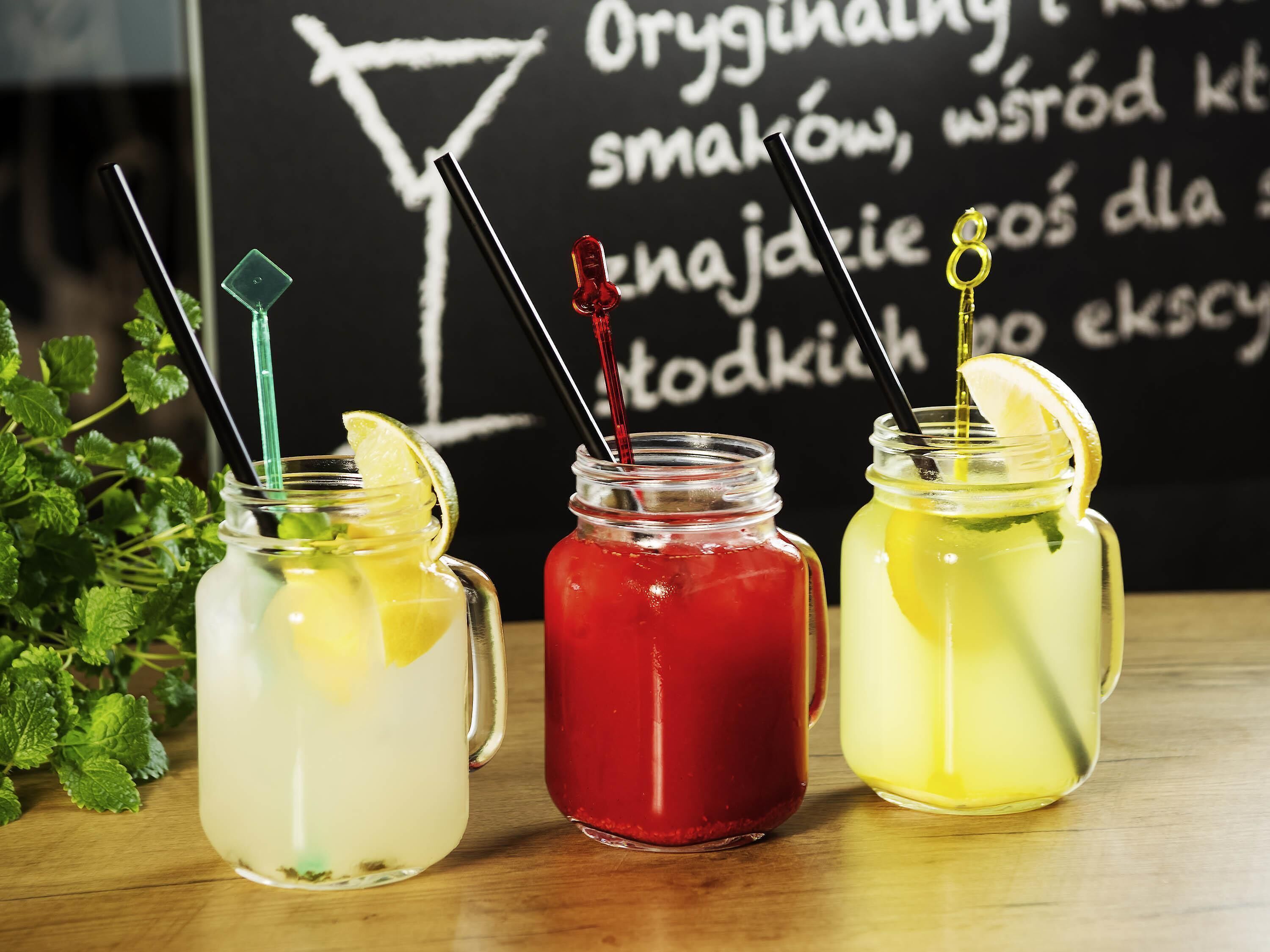 Drie glazen potten met rietjes, elk gevuld met een drankje in een andere kleur, staan op een houten oppervlak met een schoolbord op de achtergrond.