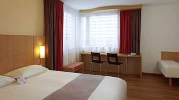 Quarto de hotel com uma cama grande, uma escrivaninha com duas cadeiras, uma mesinha e uma janela com cortinas.