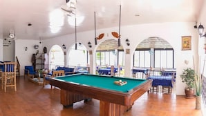 Une table de billard dans une pièce dotée de fenêtres en arc de cercle, de meubles en bois et d’une plante en pot.