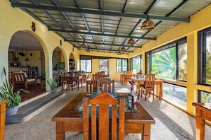 A spacious indoor area with multiple wooden tables and chairs, large windows offering views of greenery, and a ceiling with exposed beams and hanging lights. - Blue Star Dive Resort (Anda)