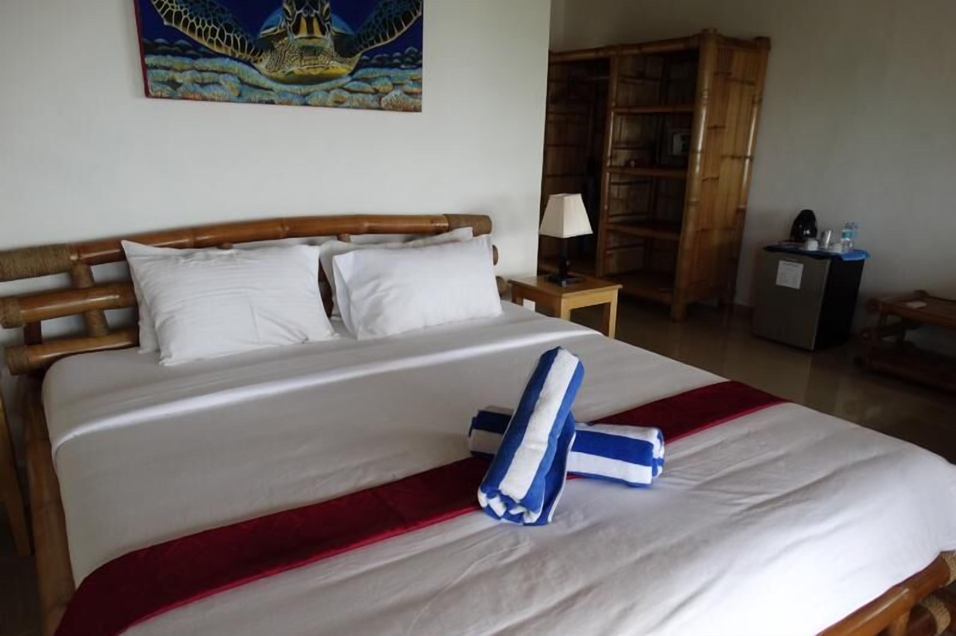 Ein ordentlich bezogenes Bett mit weißen Leinentüchern und einer Decke in blau-rot gestreiftem Muster, ein hölzerner Kopfteil, ein Nachttisch mit Lampe sowie ein Bücherregal in einem Hotelzimmer.