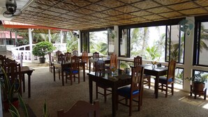 Un espace de restauration intérieur avec des tables et des chaises en bois, de grandes fenêtres offrant une vue sur les palmiers et un plafond en chaume.