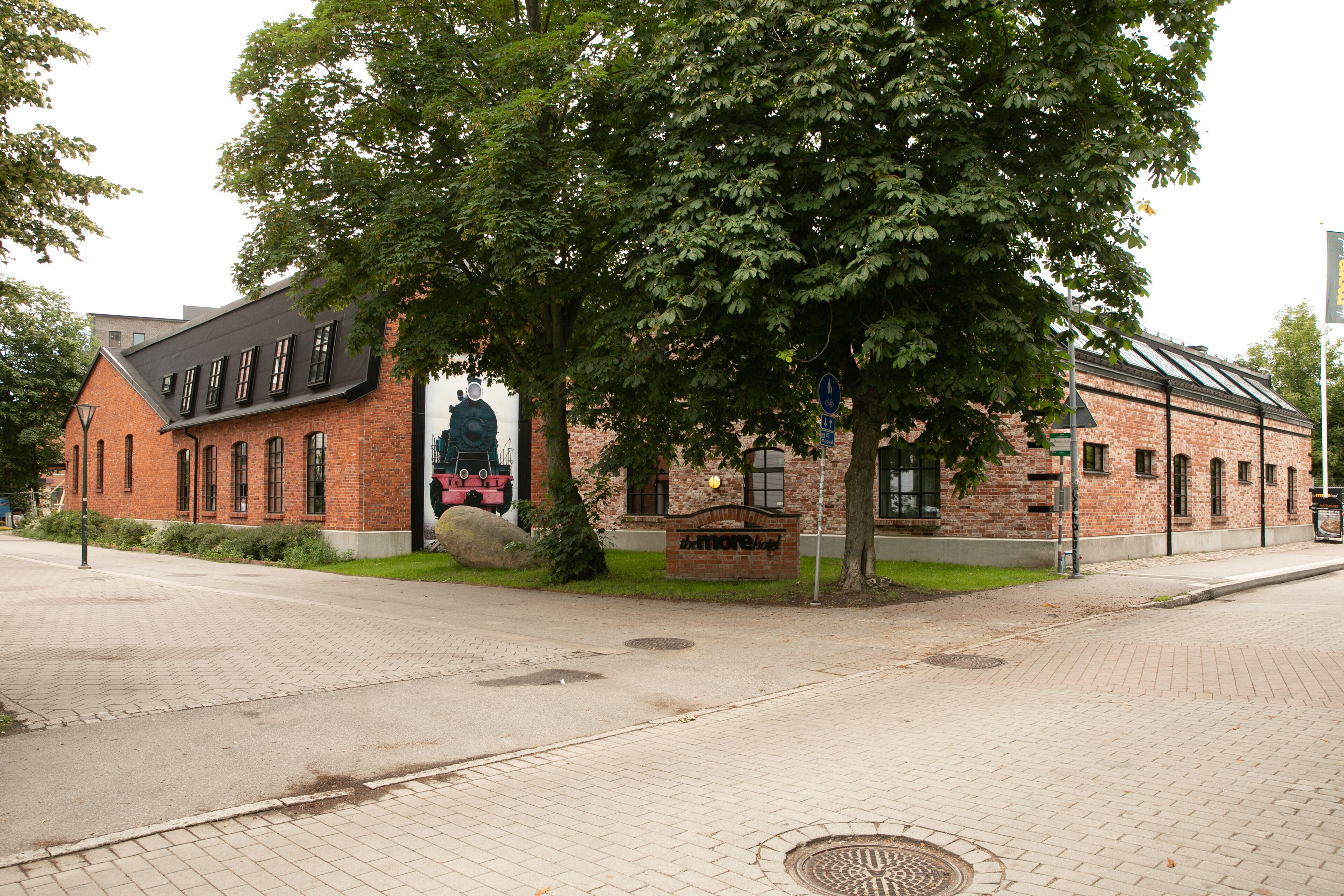 Tiilestä rakennettu rakennus, jonka edessä on suuri puu, edessä asfaltoitu jalkakäytävä ja sivulla seinämaalaus.