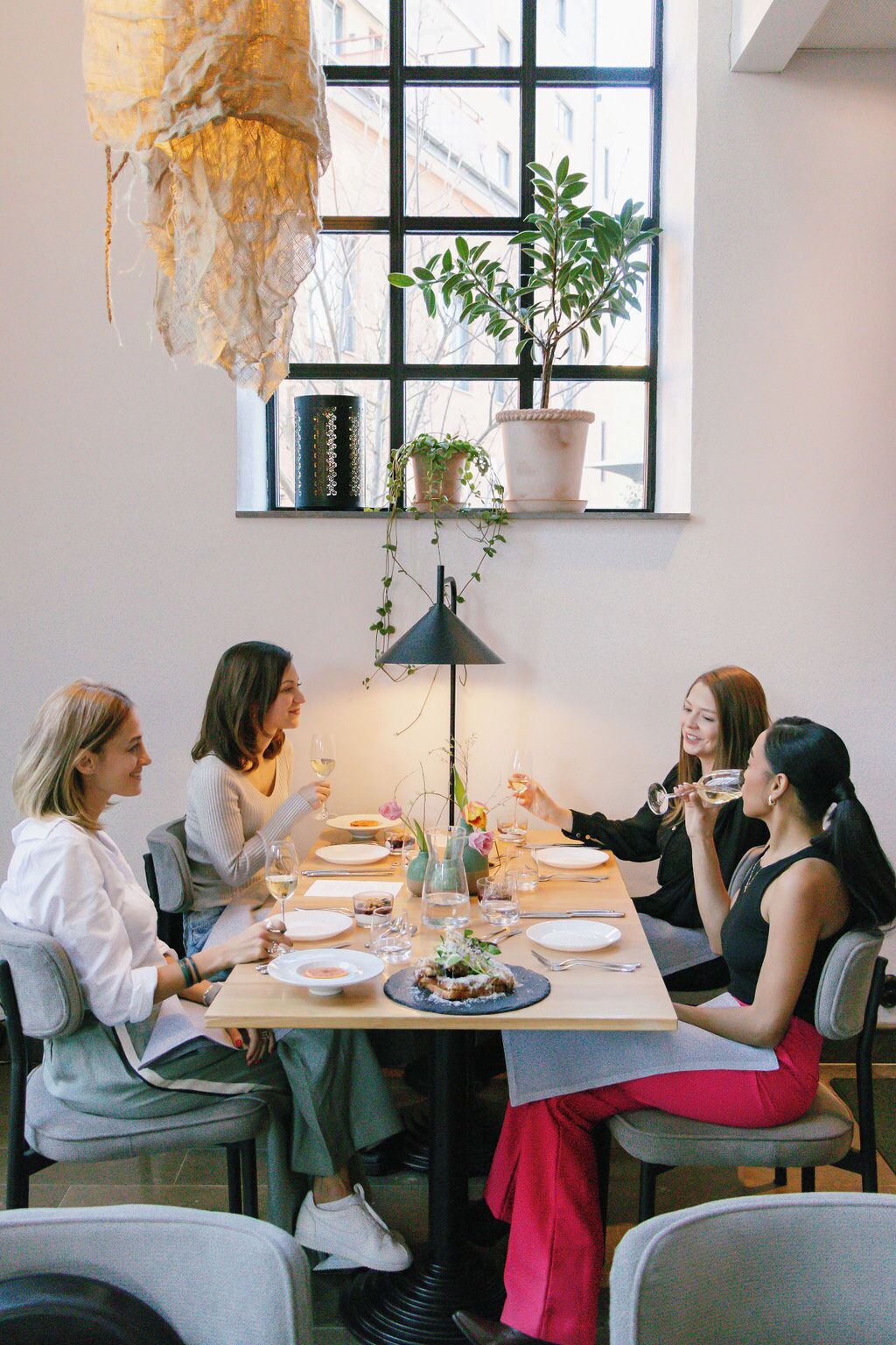 Un grupo de personas cenando en una mesa con plantas en macetas y una lámpara colgante.