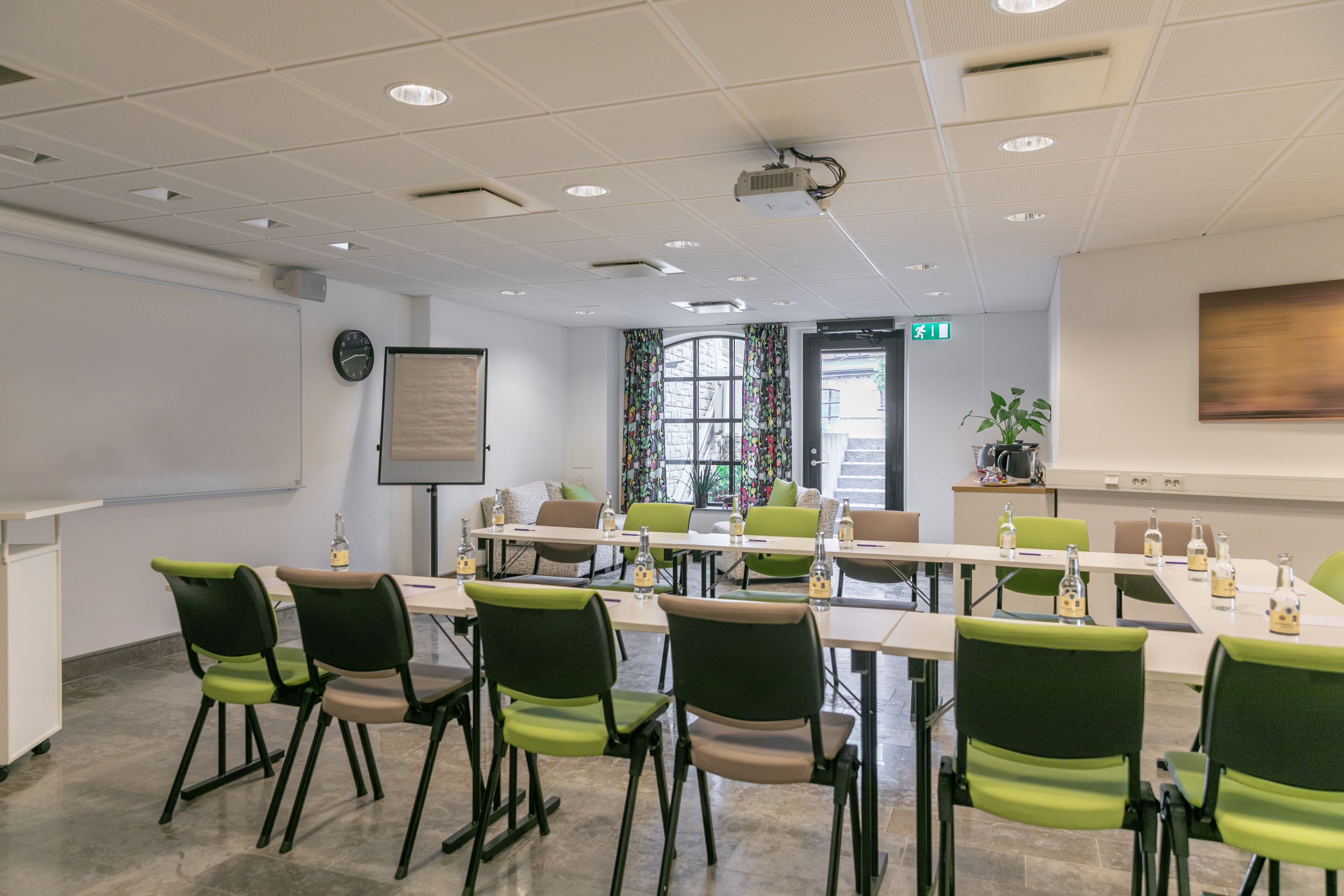 Ett konferensrum med ett långt bord, stolar, en projektor, en klocka och ett fönster med gardiner.