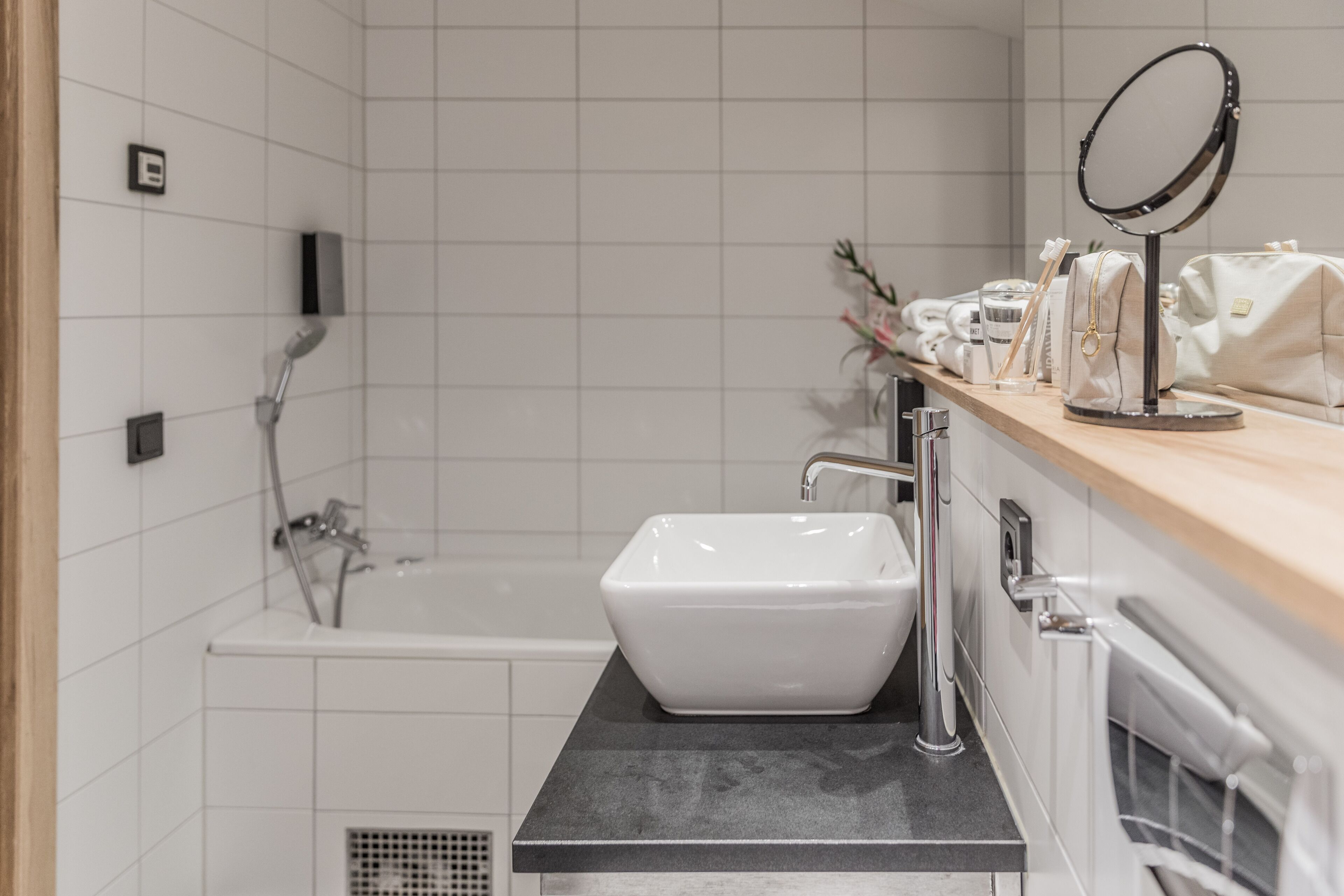 A modern bathroom with a white sink, a handheld showerhead, and a vanity with a mirror and toiletries.