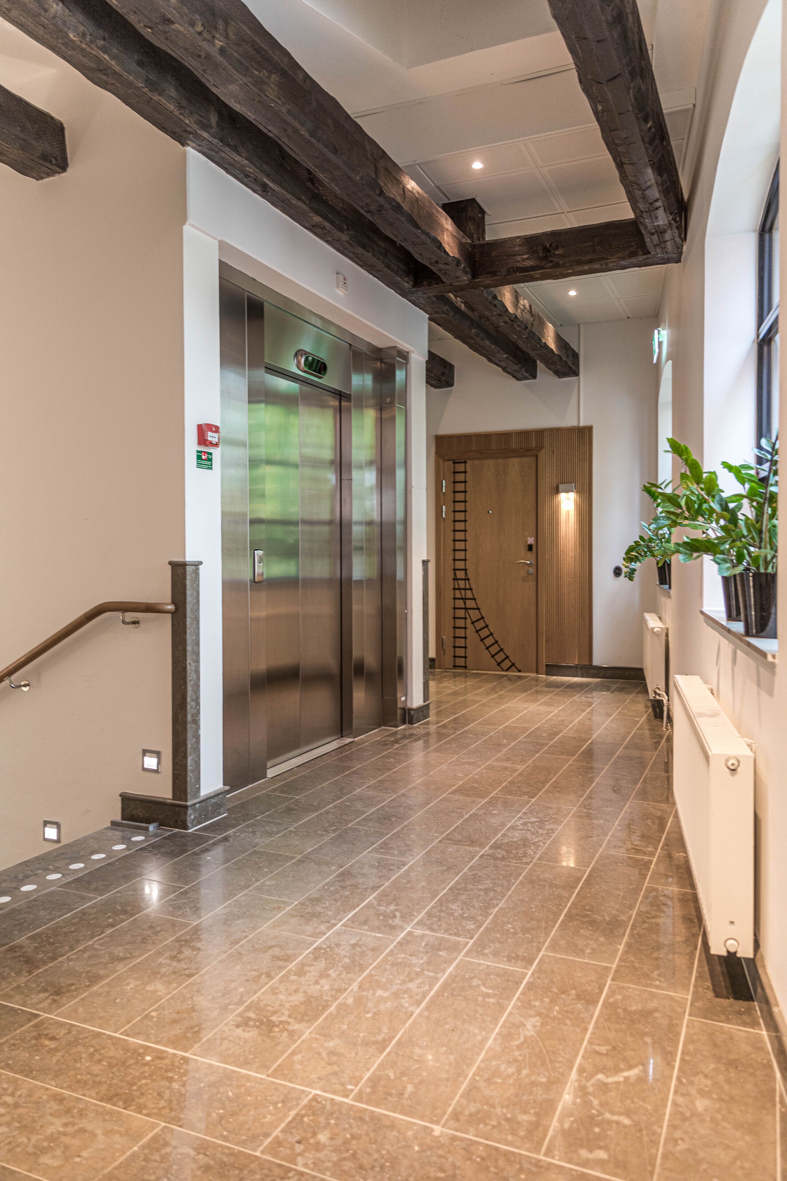 Un couloir avec un ascenseur moderne, un escalier doté d’une rampe et une plante en pot.