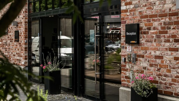 Entrada de un hotel con una recepción, un letrero negro sobre la puerta y fachada de ladrillo.