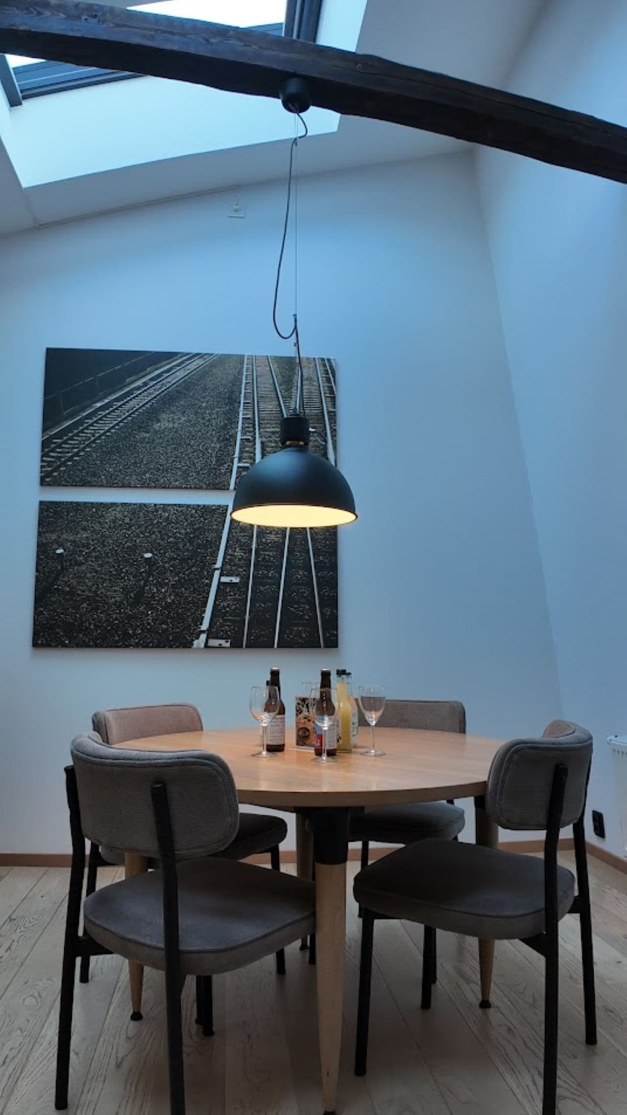 A dining area with a round table, four chairs, a black pendant light, and a large railway track artwork on the wall.