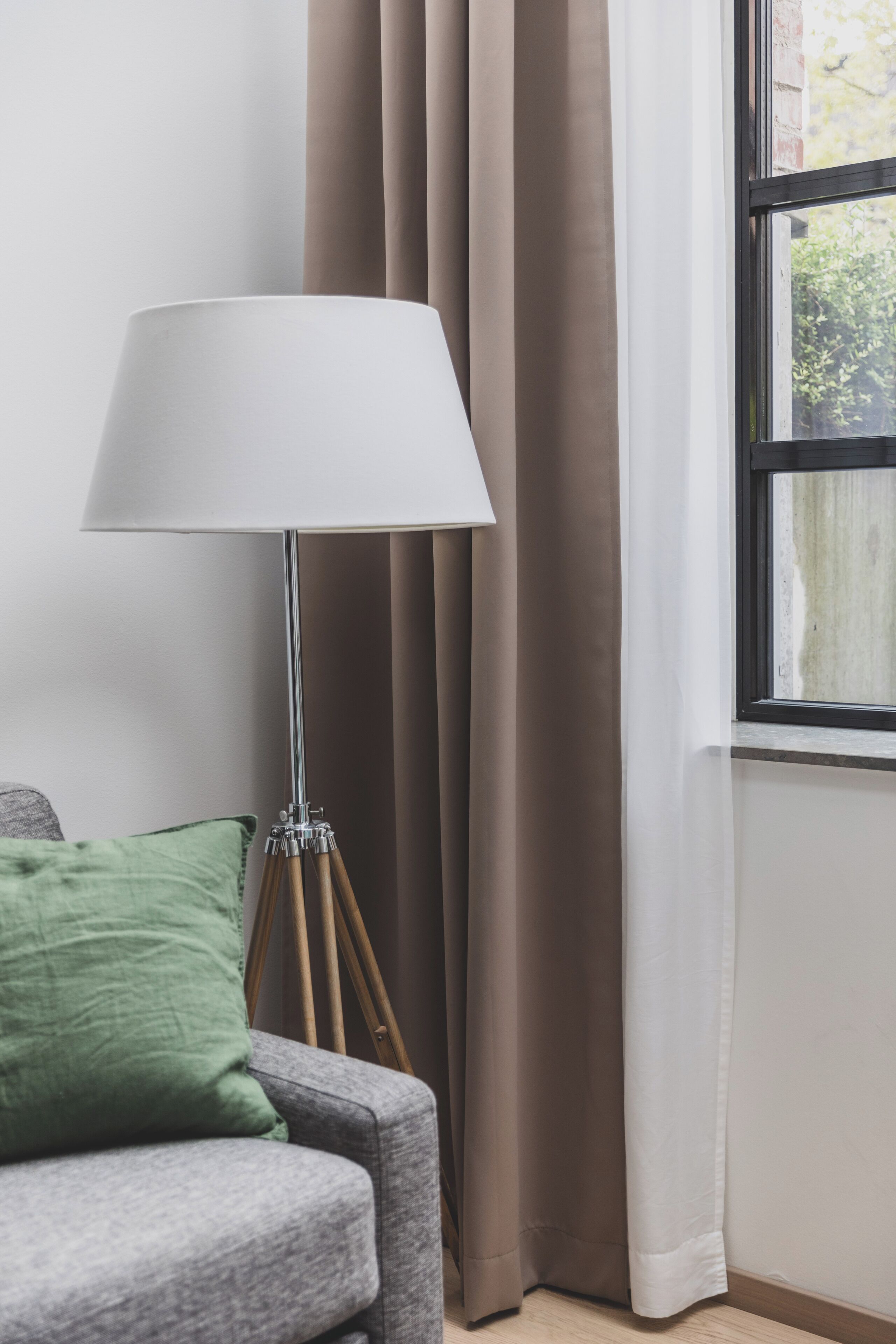 A room with a grey sofa, a green pillow, a floor lamp with a white shade, and a window with curtains.