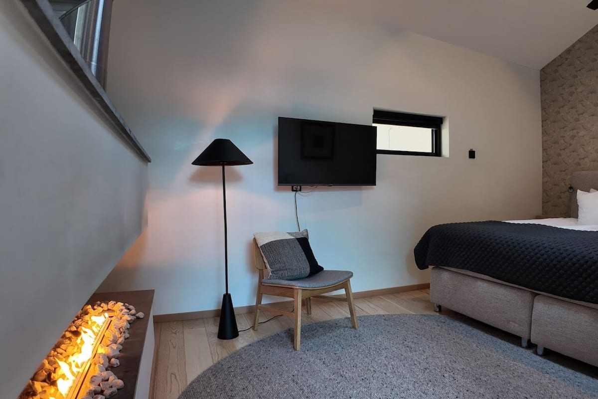 A bedroom with a bed, a chair, a TV mounted on the wall, and a fireplace.