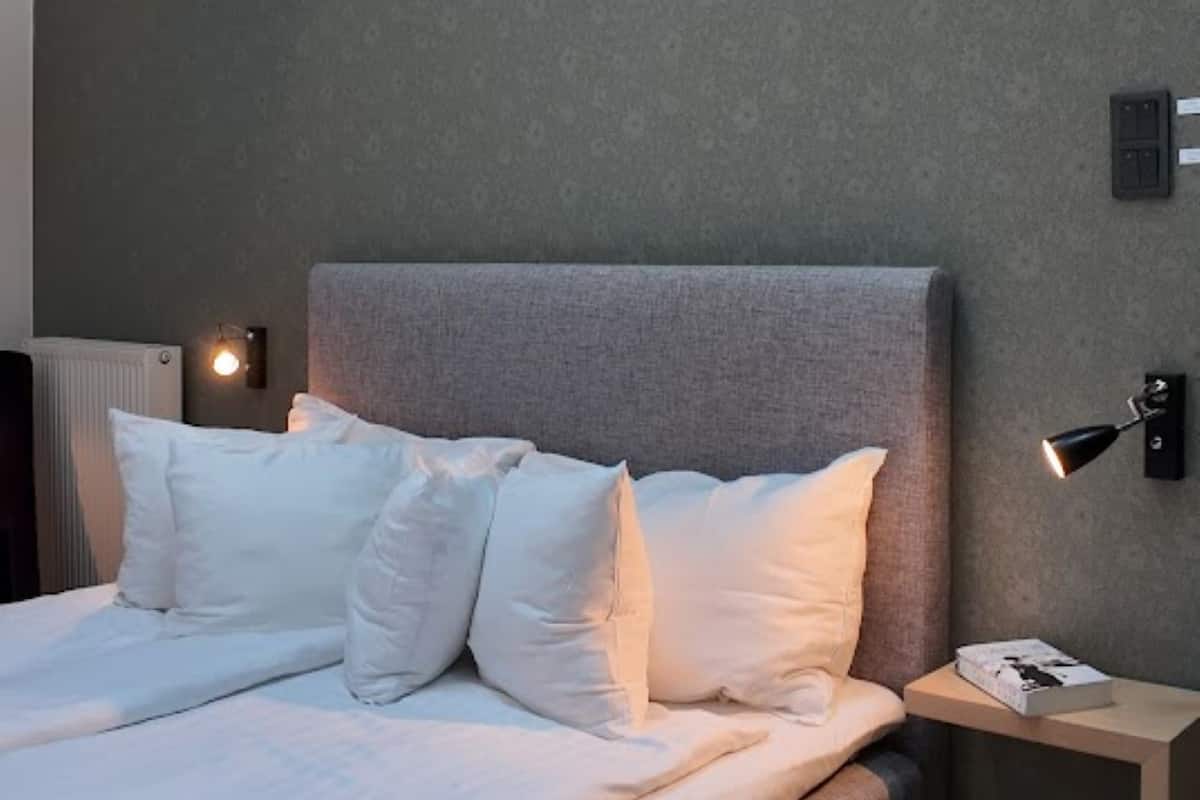 A modern bedroom with a grey upholstered bed, white pillows, a bedside table with a lamp, and a wall-mounted light fixture.