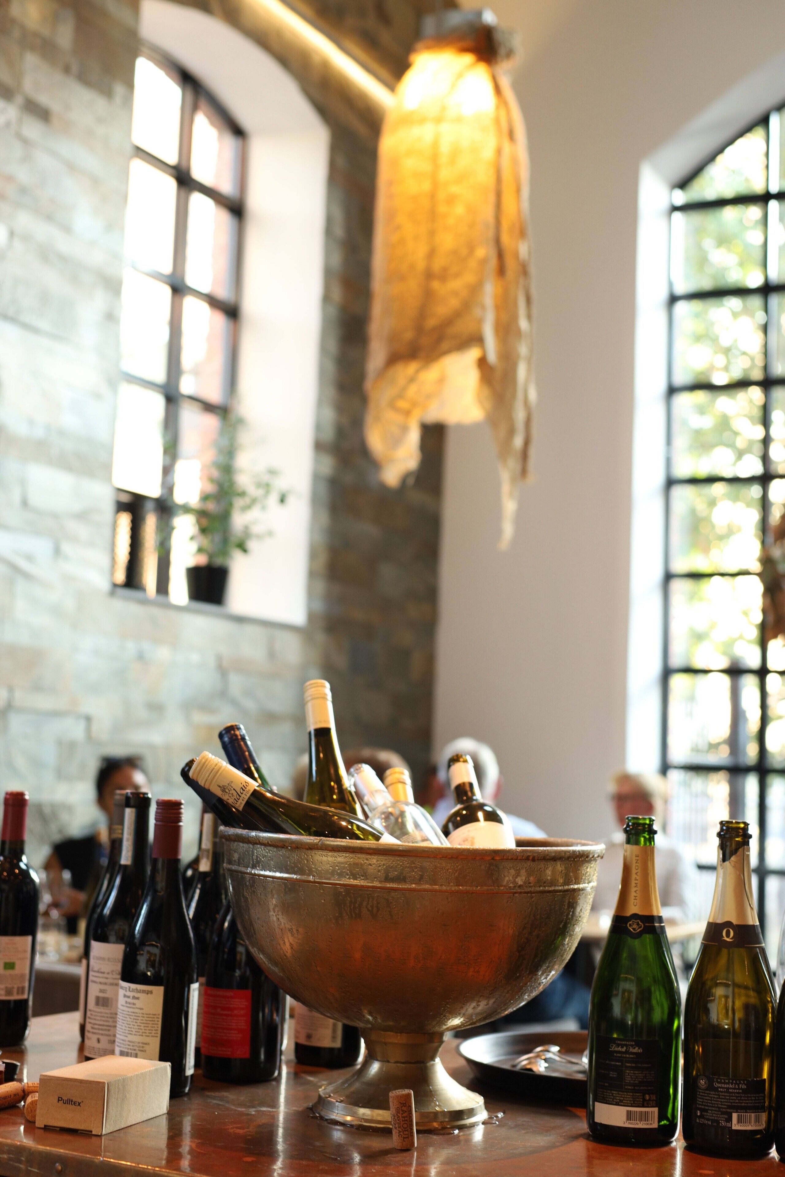 A bar area with a metal bowl filled with bottles, a hanging light fixture, and a stone wall with windows.