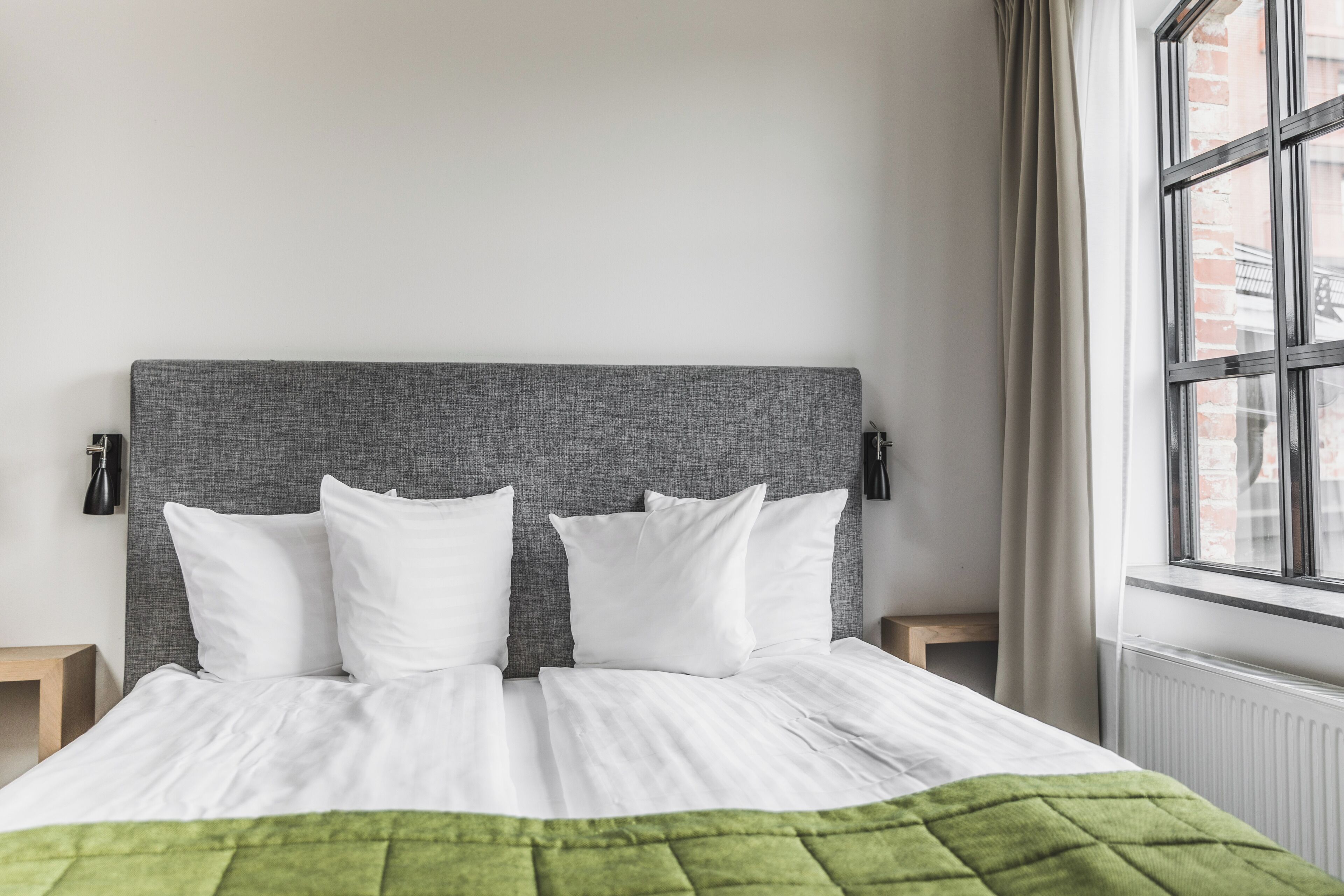Una cama bien hecha con ropa de cama blanca y un edredón verde, un cabecero gris y una ventana con cortinas.