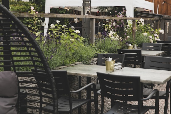 An outdoor patio with tables and chairs, surrounded by lush greenery and flowering plants.