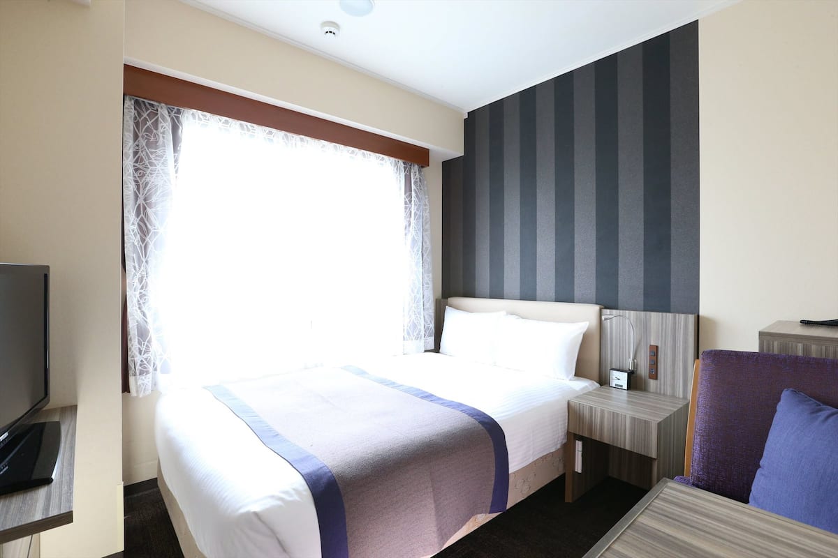 A hotel room with a large bed, a bedside table with a lamp, a TV, and a view of the outdoors through a window with curtains.