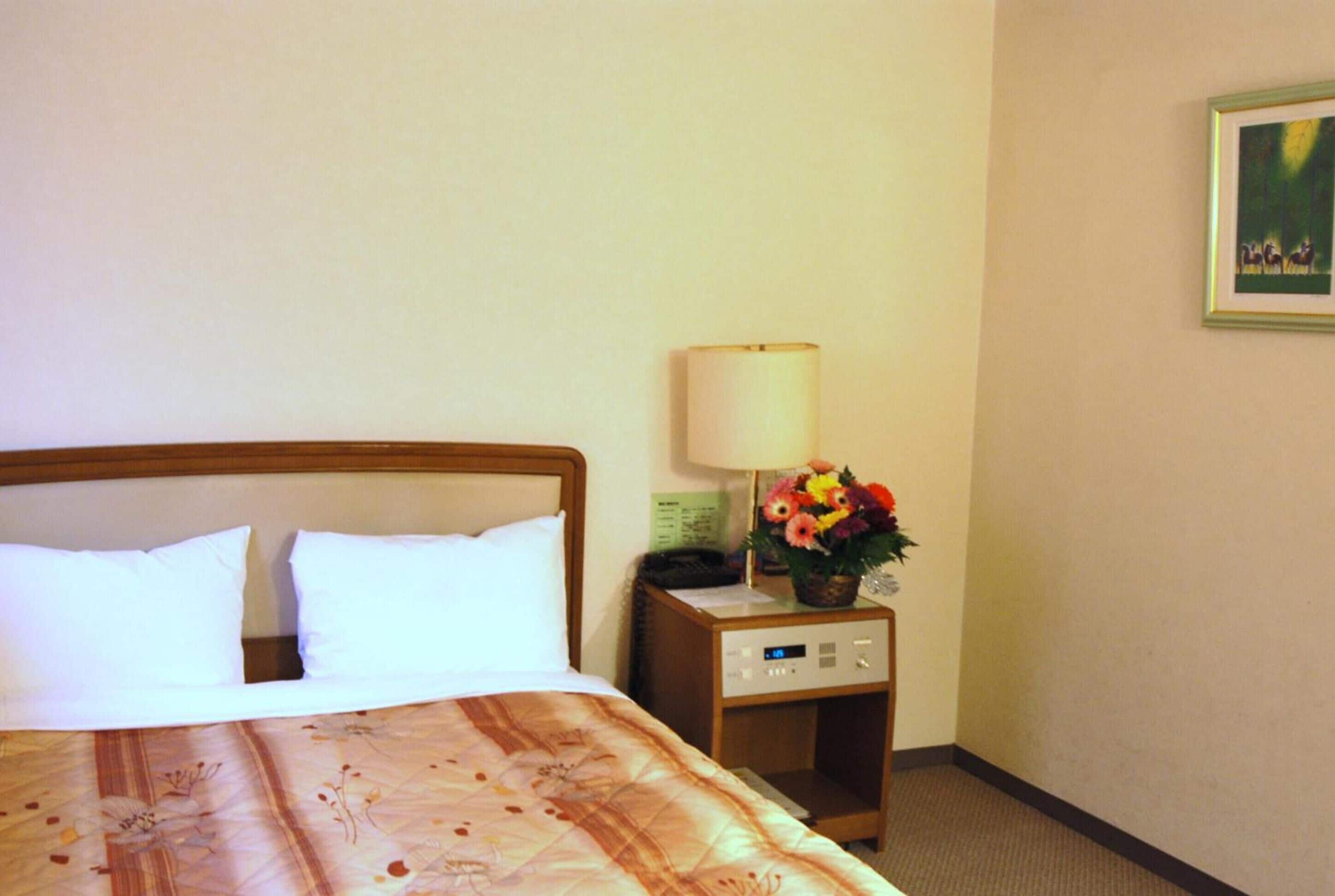 A hotel room with a bed, a nightstand with a lamp and flowers, and a painting on the wall.