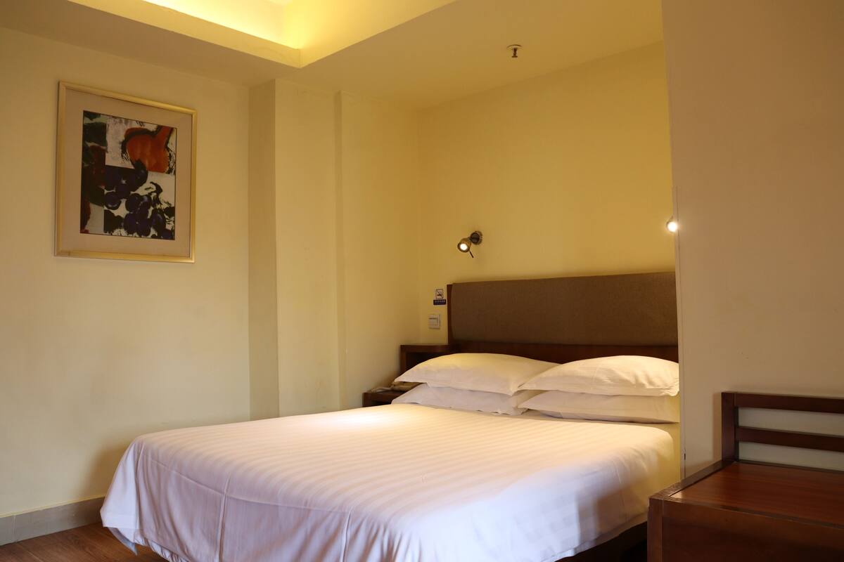 Een hotelkamer met een enkel bed, een houten hoofdbord, een nachtkastje en een lijst met een abstract schilderij aan de muur.