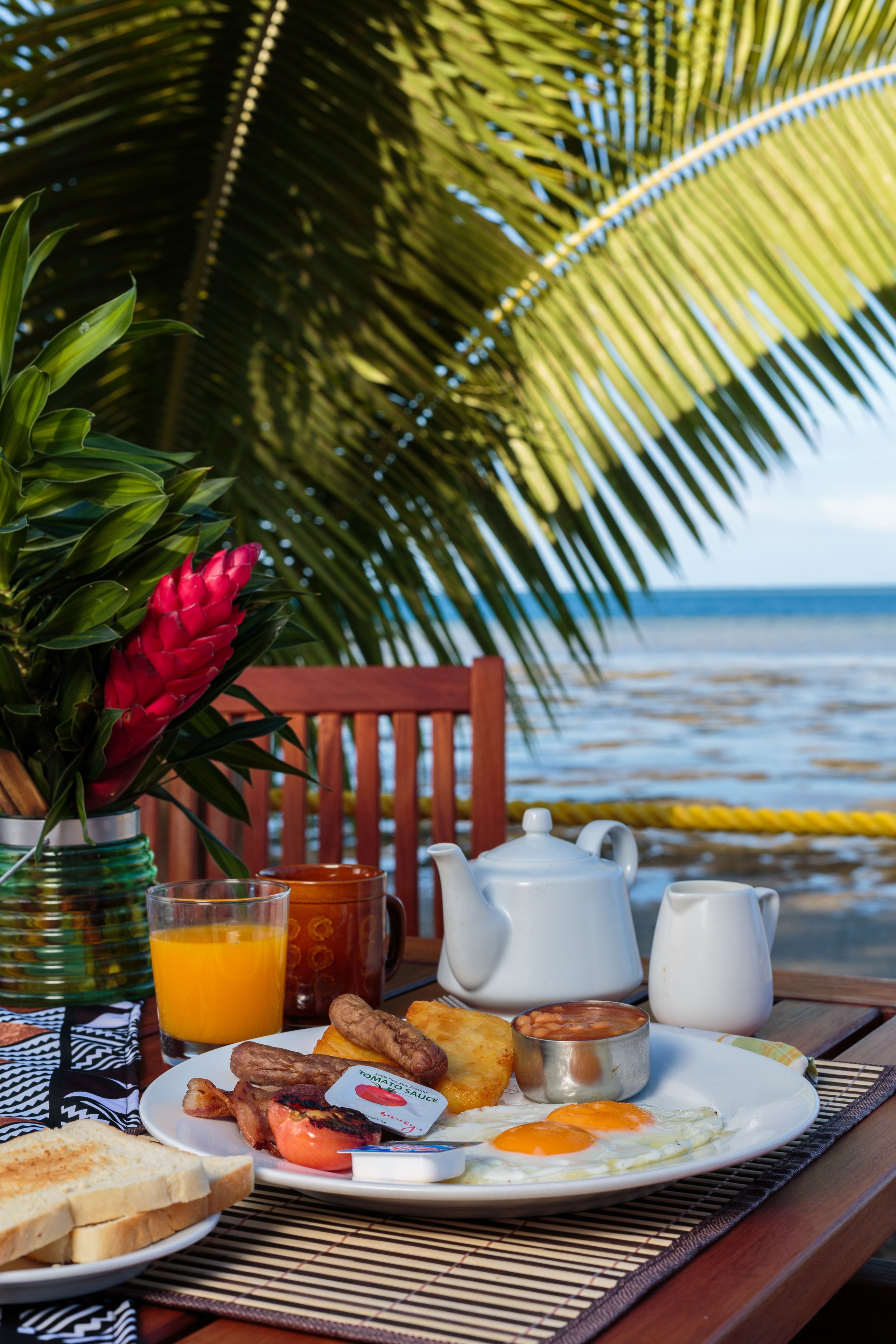 A breakfast table with a plate of eggs, toast, and beans, a glass of orange juice, and a pitcher of milk, set against a backdrop of a palm tree and ocean view.