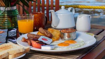 Una mesa de desayuno con un plato de huevos, tostadas y frijoles, un vaso de jugo de naranja y una jarra de leche, con un palmera y vista al océano de fondo.