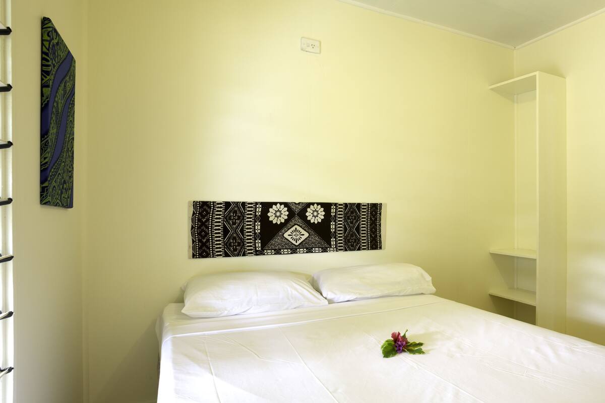 A neatly made bed with white linens and a decorative pillow, a wall-mounted tapestry with a geometric pattern, and a wardrobe with a mirror and a patterned curtain.