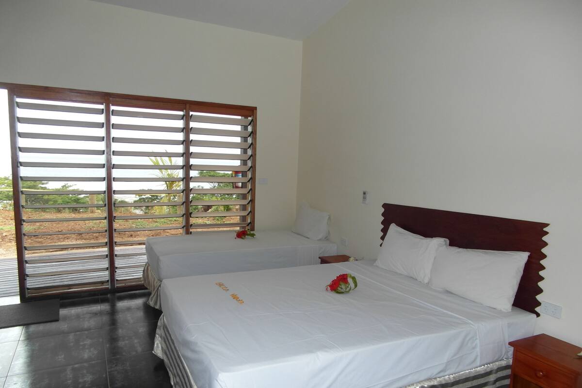 A hotel room with two beds, a wooden headboard, a small bedside table, and a view through a sliding door.