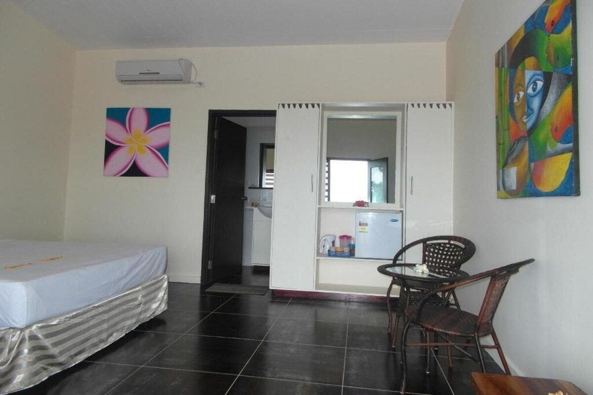 A room with a bed, a chair, a table, an air conditioning unit, and a door leading to a kitchen area.