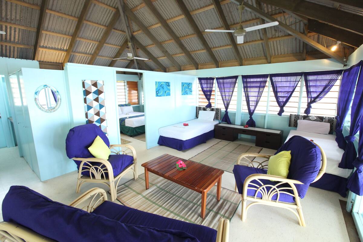 A room with a high ceiling, wooden beams, a bed with a blue bedspread, two chairs with blue cushions, a wooden table, and a window with purple curtains.