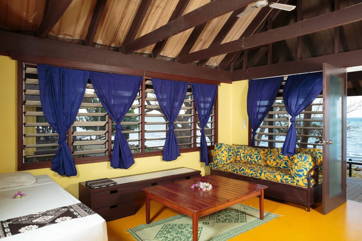 A room with a wooden ceiling, a sofa with a floral pattern, a bed with a white and black bedspread, and a window with blue curtains.