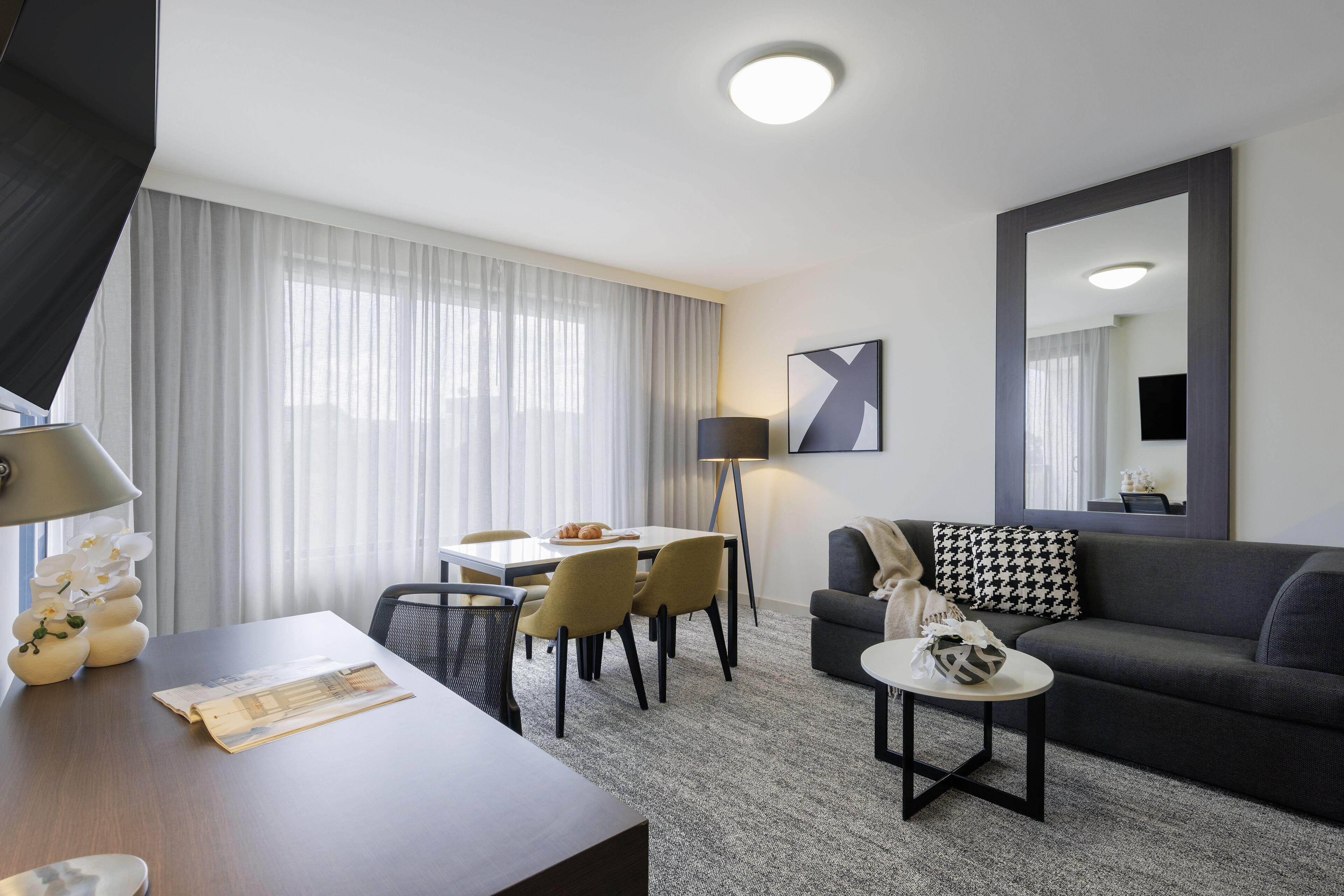 A modern hotel room with a dining area, a sofa, a coffee table, and a large mirror.