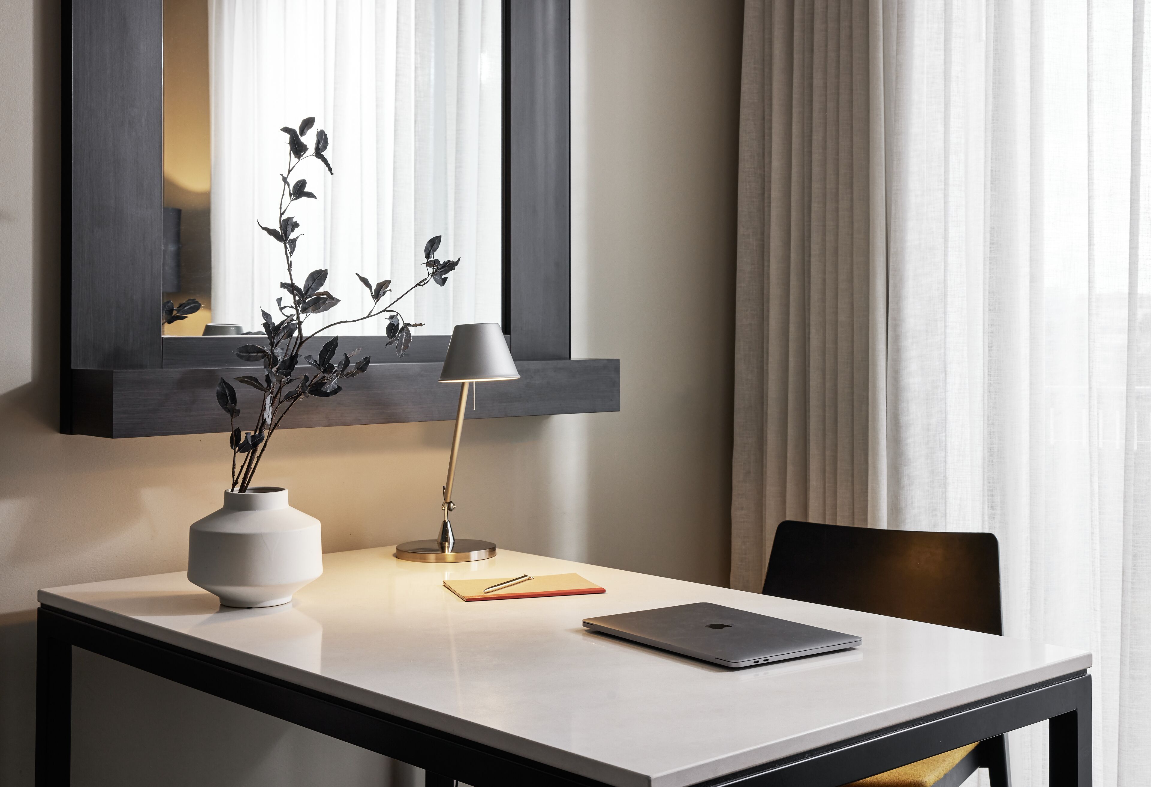 Una habitación moderna con un escritorio, una computadora portátil, una lámpara, un jarrón con plantas y un espejo.