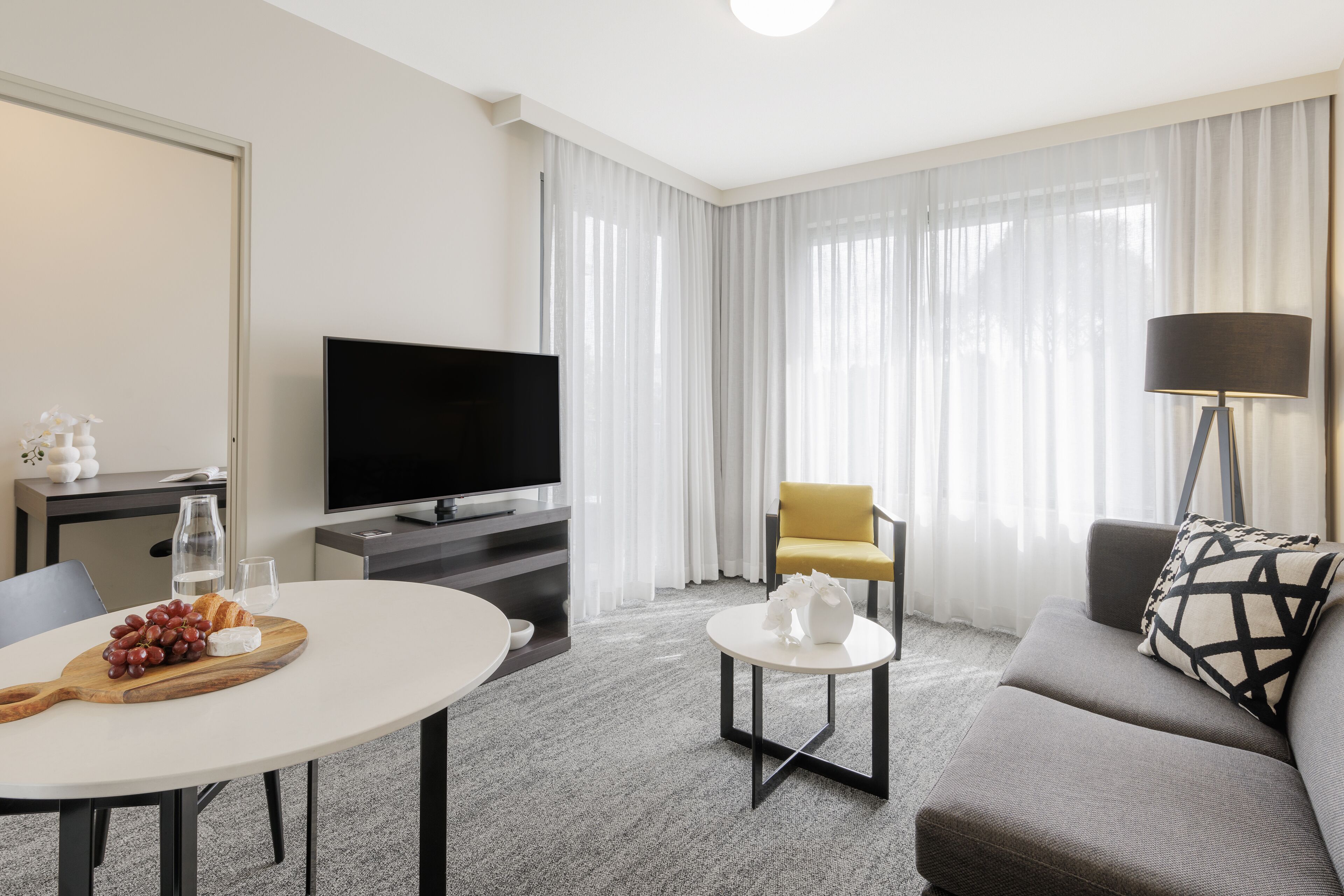 A modern hotel room with a flat-screen TV, a dining table with a fruit platter, a sofa, a yellow armchair, and a lamp.