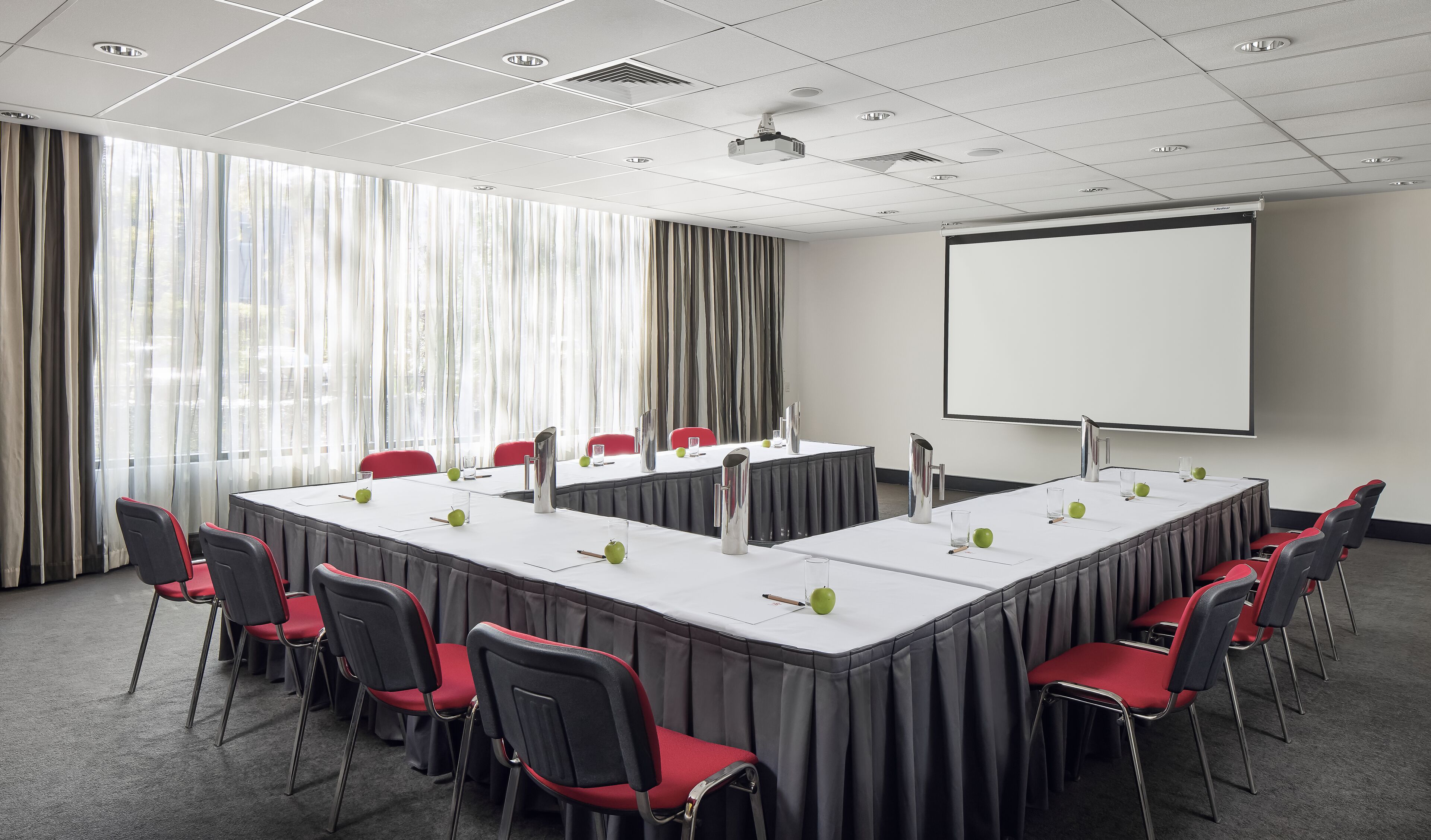 A conference room with a long table, red chairs, and a projector screen.