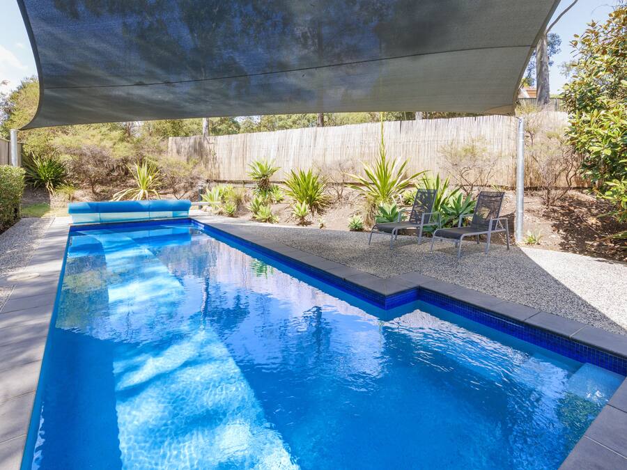 A rectangular swimming pool with a blue tiled edge, surrounded by a concrete deck. There are lounge chairs and a shaded area overhead.