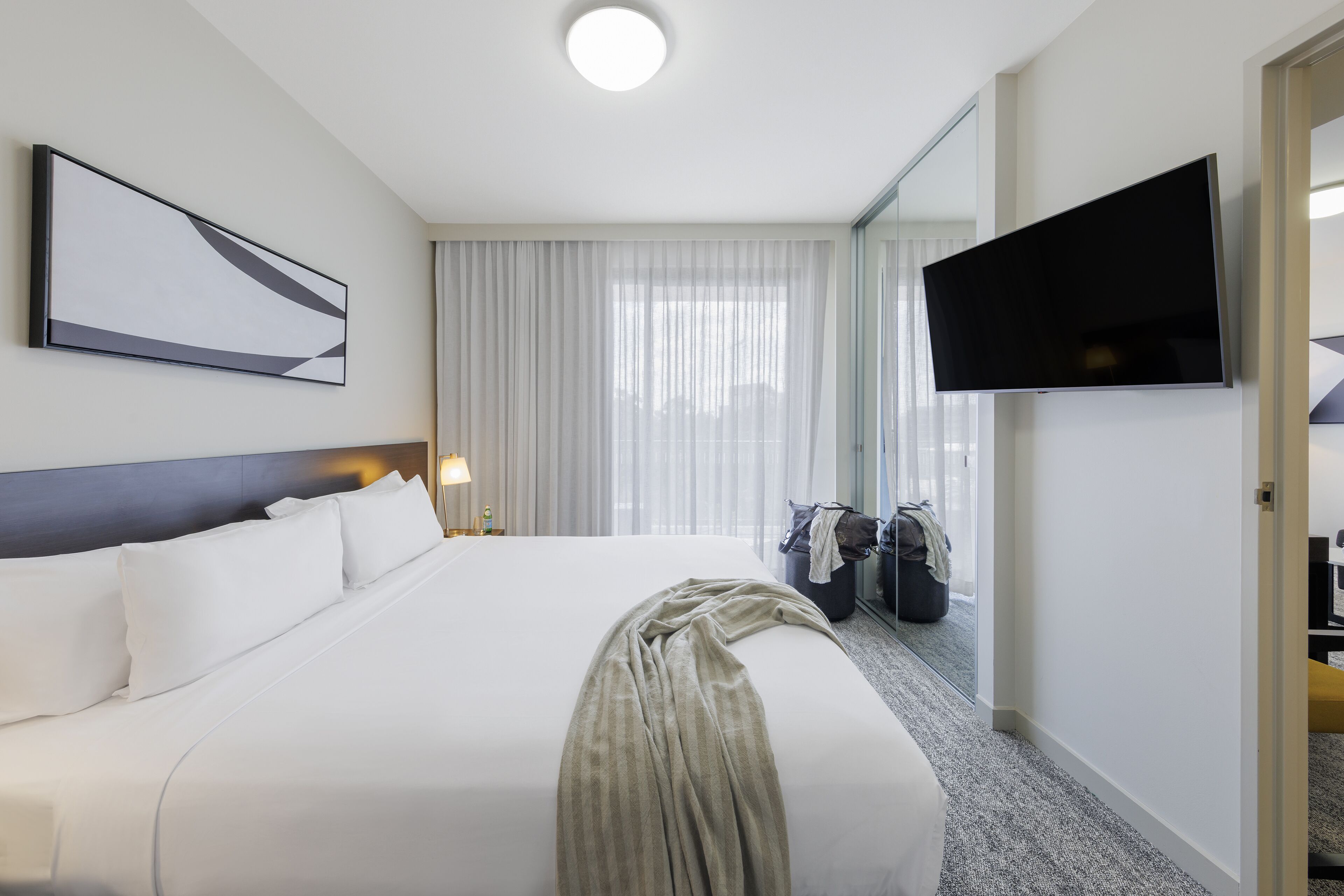 A hotel room with a large bed, a flat-screen TV mounted on the wall, a window with sheer curtains, and a coat rack with a jacket.