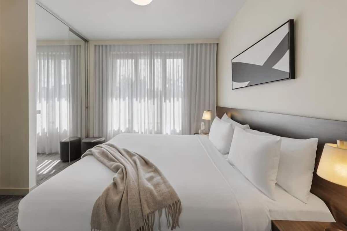 A hotel room with a large bed, a bedside table with a lamp, a view of a window with sheer curtains, and a modern abstract painting on the wall.