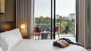 A hotel room with a bed, a nightstand with a lamp, a table with chairs, and a balcony with a view of trees and buildings. - Punthill Norwest (Norwest)