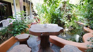 Une table ronde en bois, sur laquelle sont posés un verre et une tasse, entourée de tabourets assortis, est placée sur un sol de gravier dans un jardin luxuriant agrémenté d’un bassin.