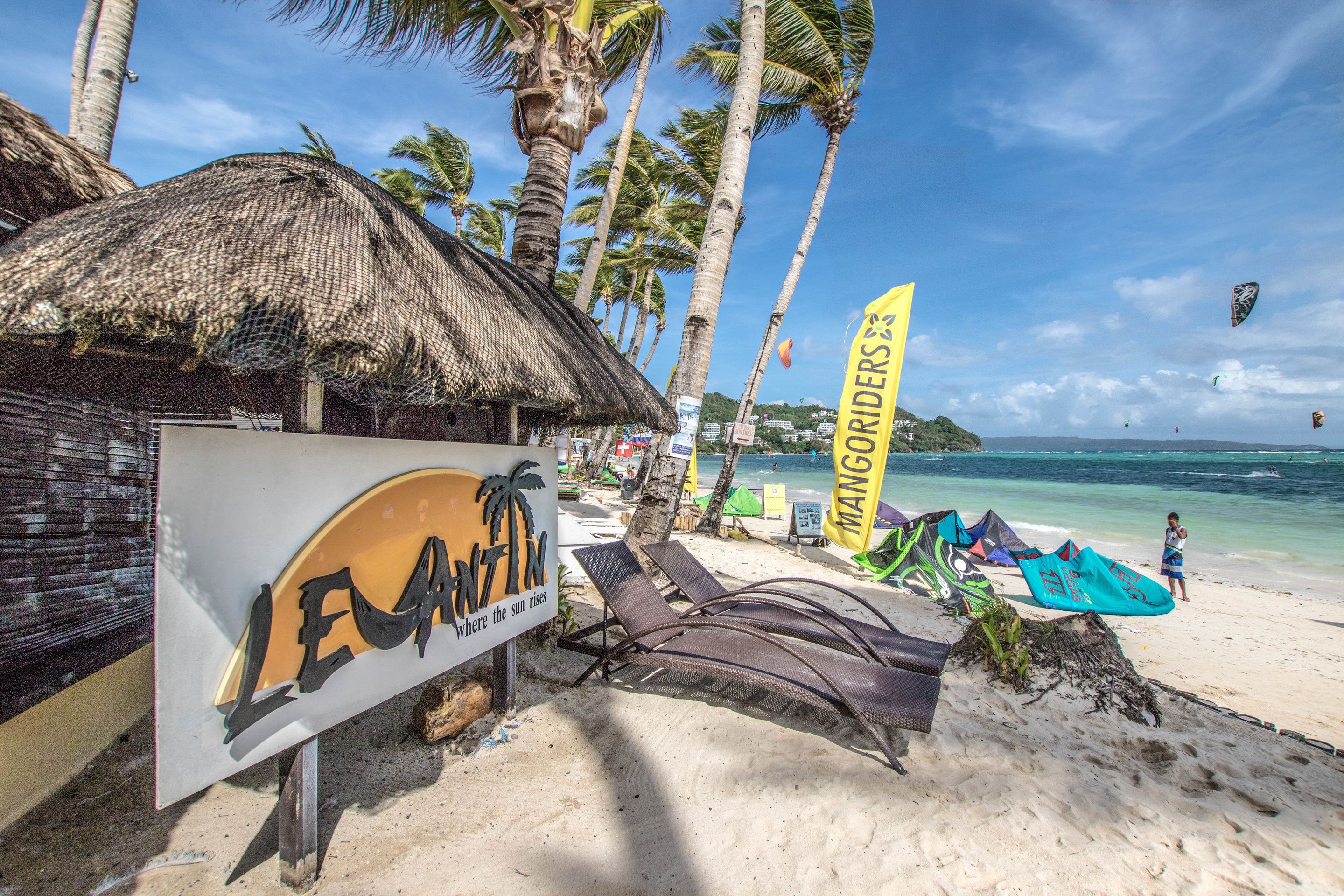 A beach with a thatched-roof hut, lounge chairs, and a sign for 'LeAntin'.