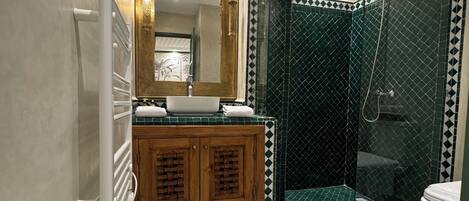 Un baño con lavamanos blanco, armarios de madera, un espejo grande y una zona de ducha con azulejos estampados en verde y blanco.