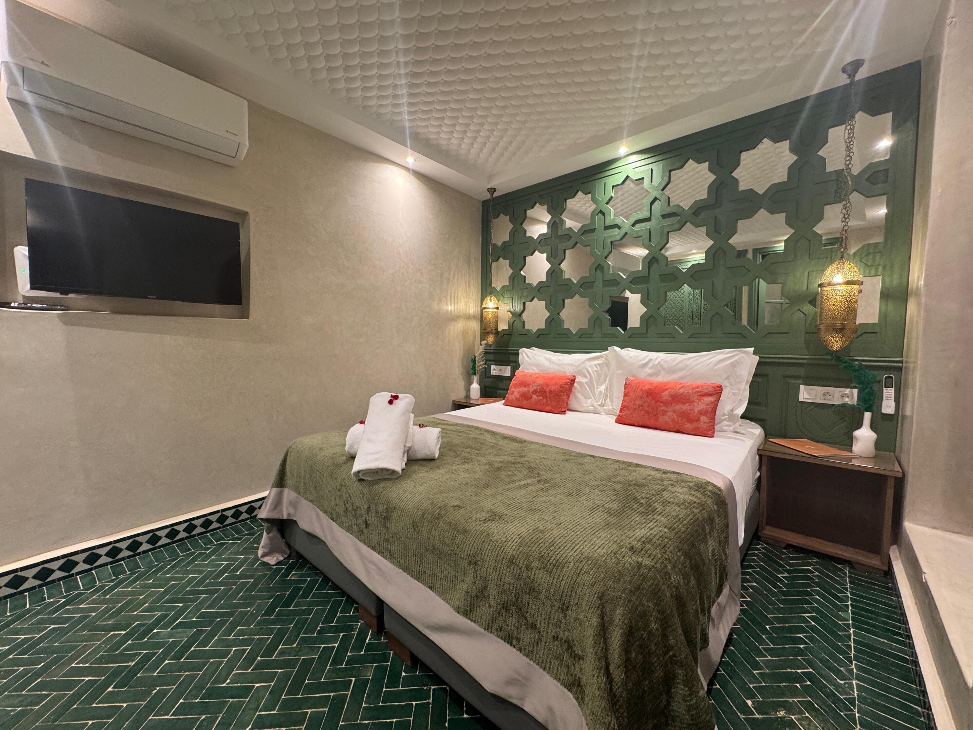A hotel room with a green herringbone floor, a bed with white and red pillows, a green throw blanket, and a wall-mounted TV.