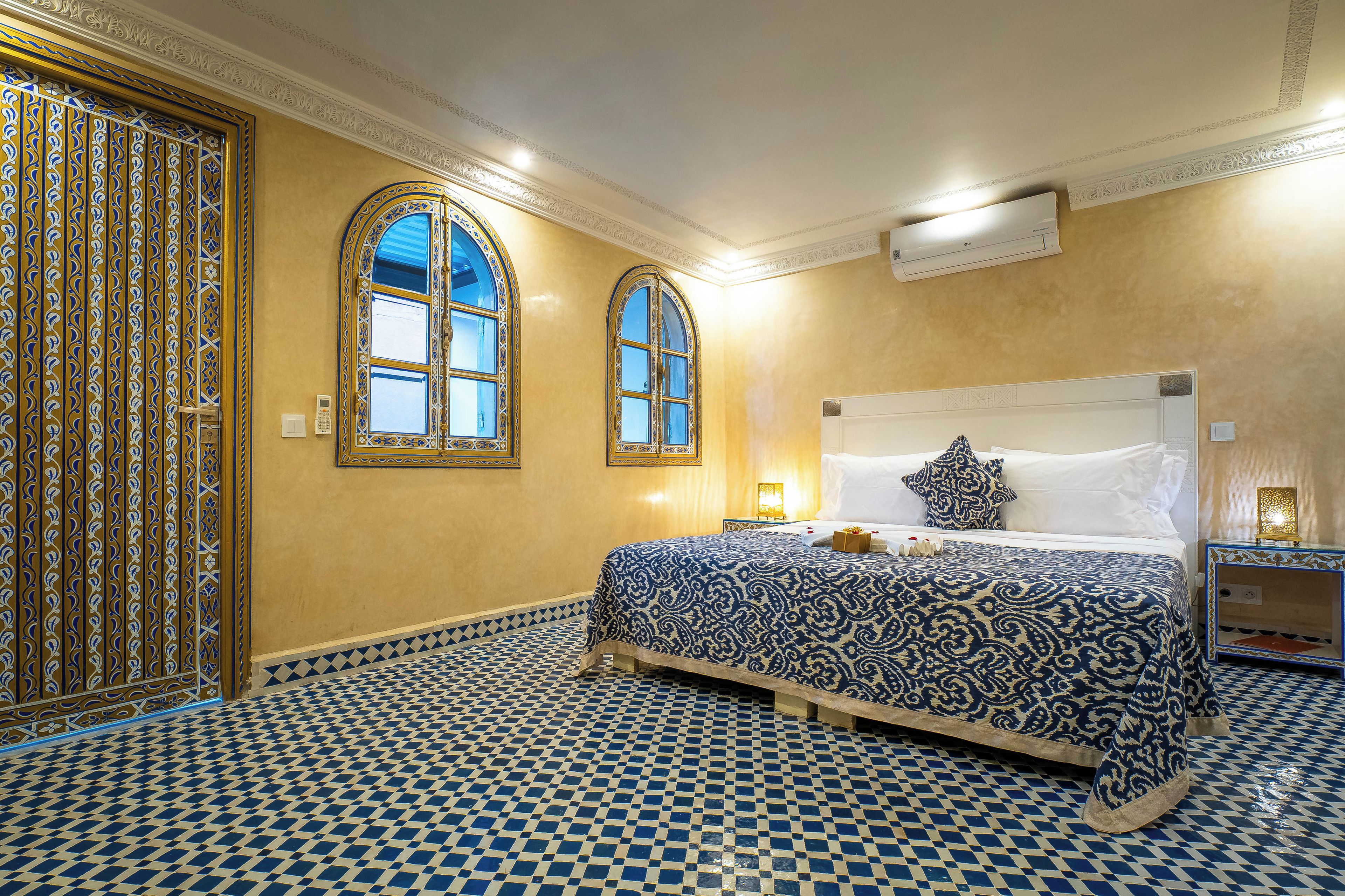 A hotel room with a patterned floor, a bed with a patterned bedspread, two arched windows, and a decorative wall panel.