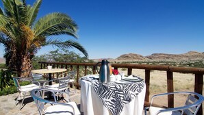 An outdoor dining area with a table set for two, surrounded by metal chairs, a zebra-patterned tablecloth, and a view of distant hills.