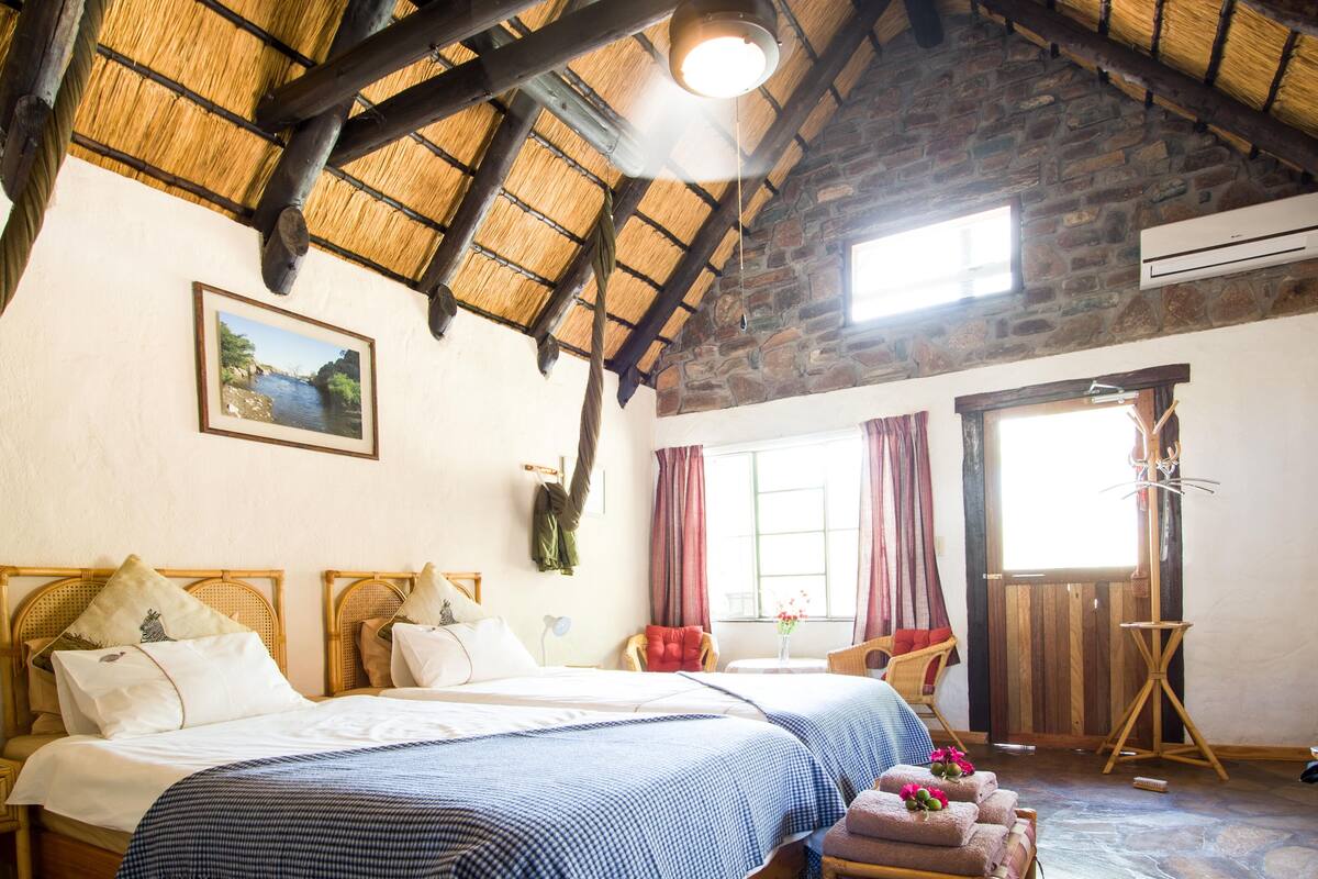 A room with two beds, a wooden ceiling, a stone wall, a painting, a window, and a chair.