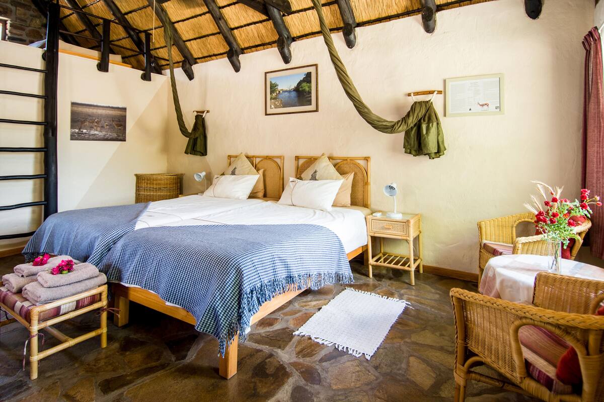 A cozy bedroom with a stone floor, a bed with a blue checkered bedspread, a wicker chair, a small table with a vase of flowers, and a wooden ladder.